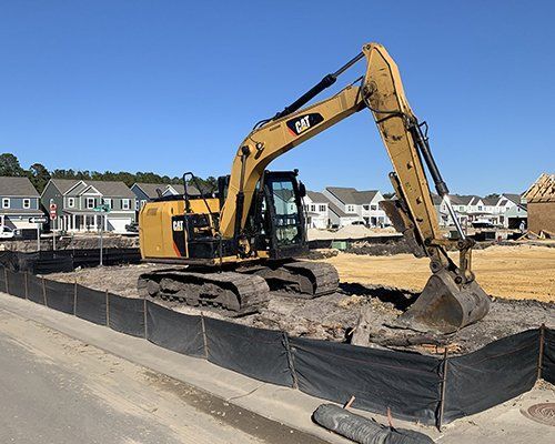 Yellow excavator digging on a construction site, black barrier, houses in the background.