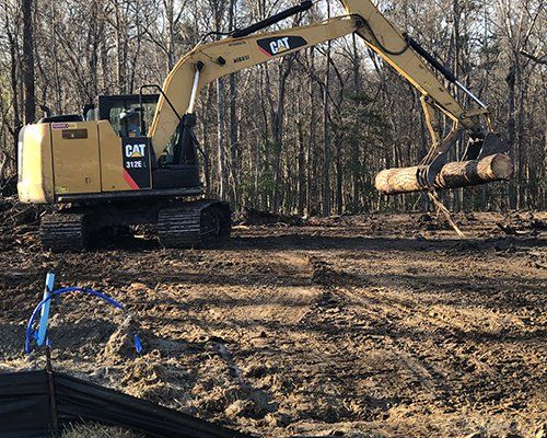 A Caterpillar excavator lifts a log in a muddy clearing, surrounded by trees.