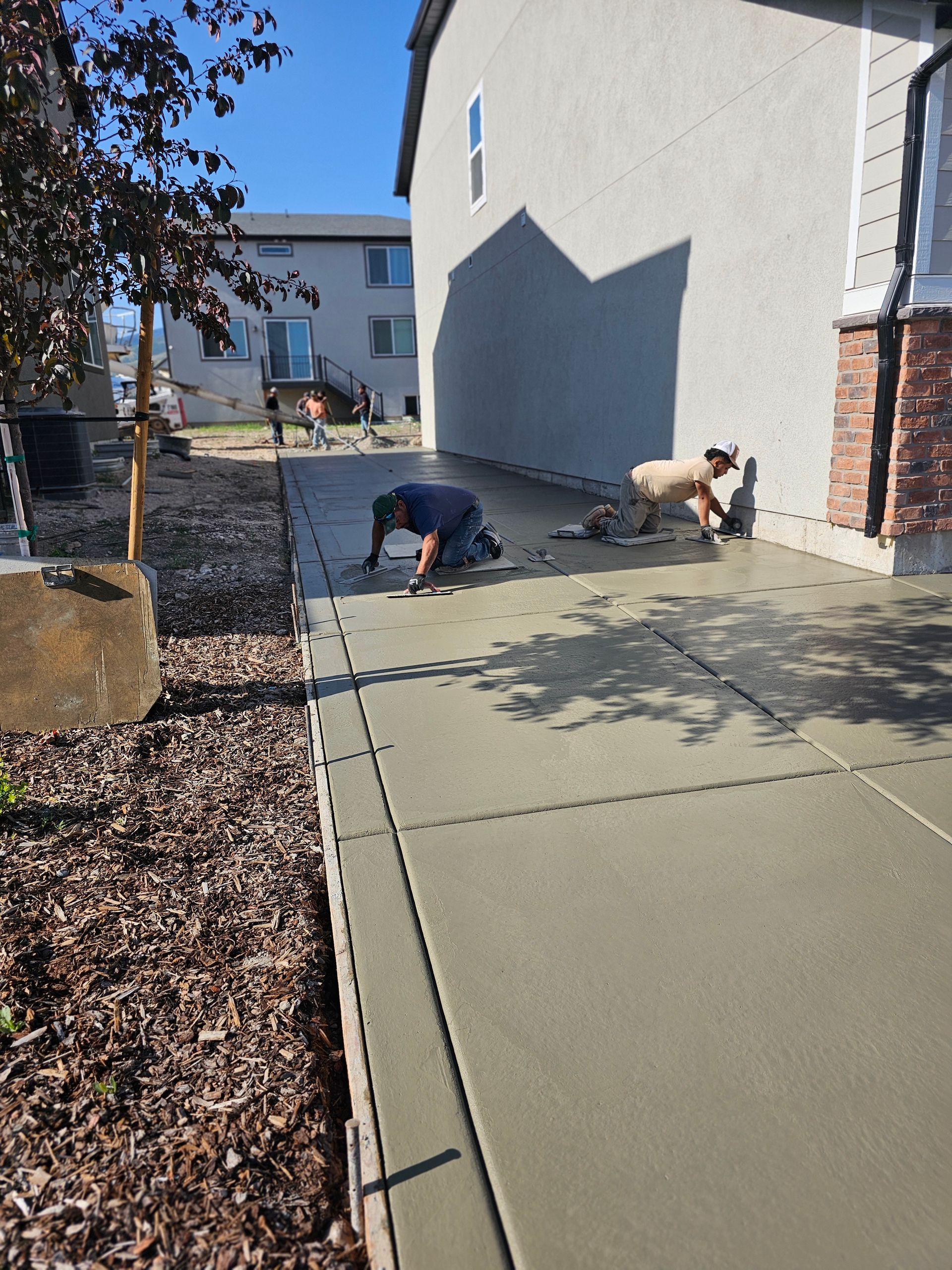 Two workers smooth a freshly poured concrete walkway alongside a house on a sunny day.