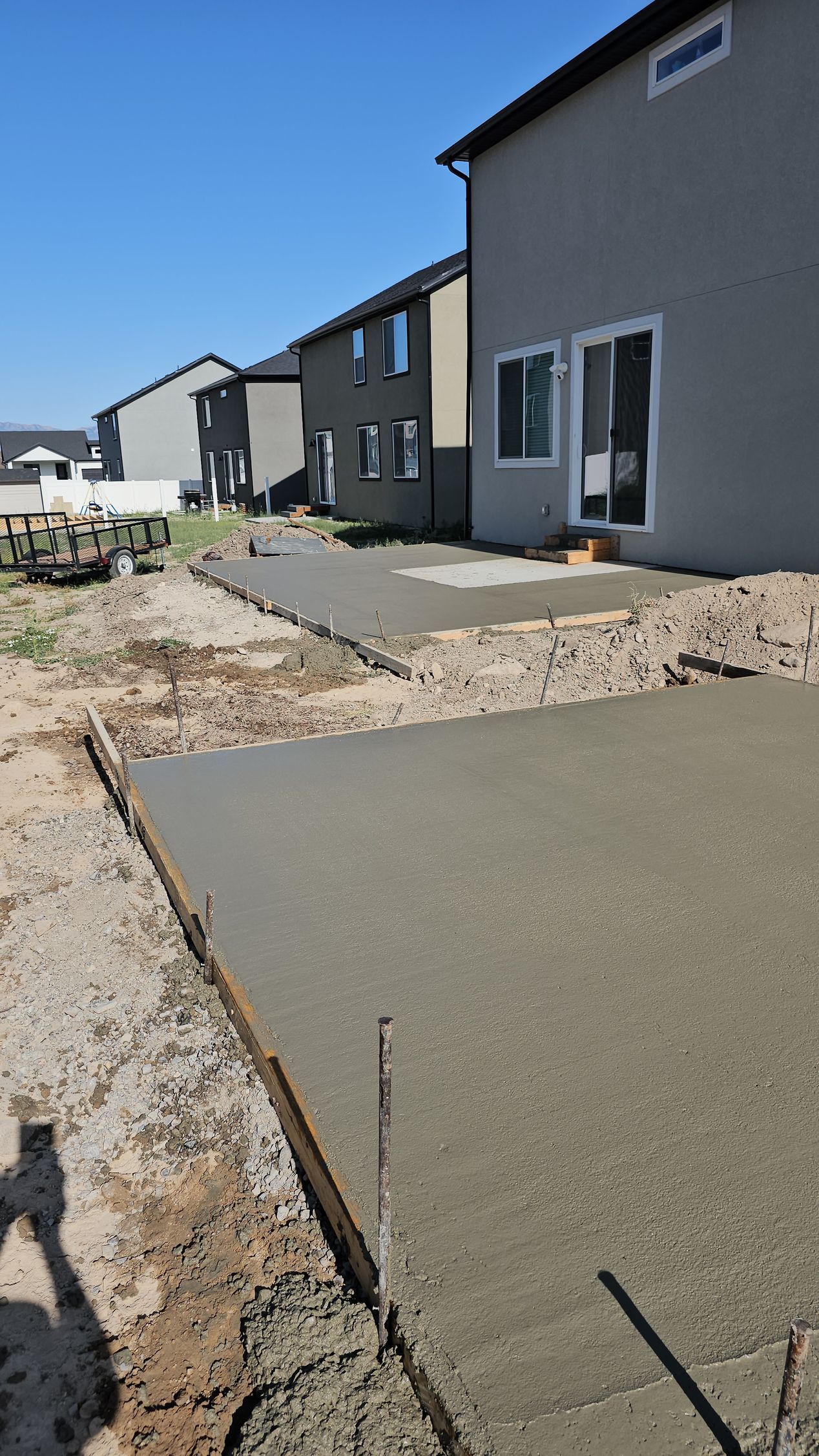 Freshly poured concrete patio slabs in the backyard of a residential house under a clear blue sky.