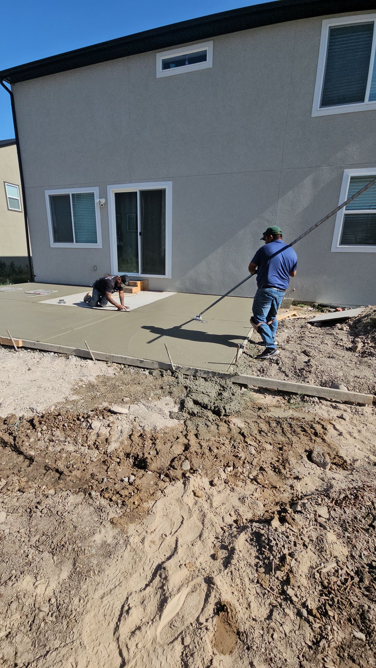 Two construction workers finish smoothing freshly poured concrete for a new patio beside a two-story house.