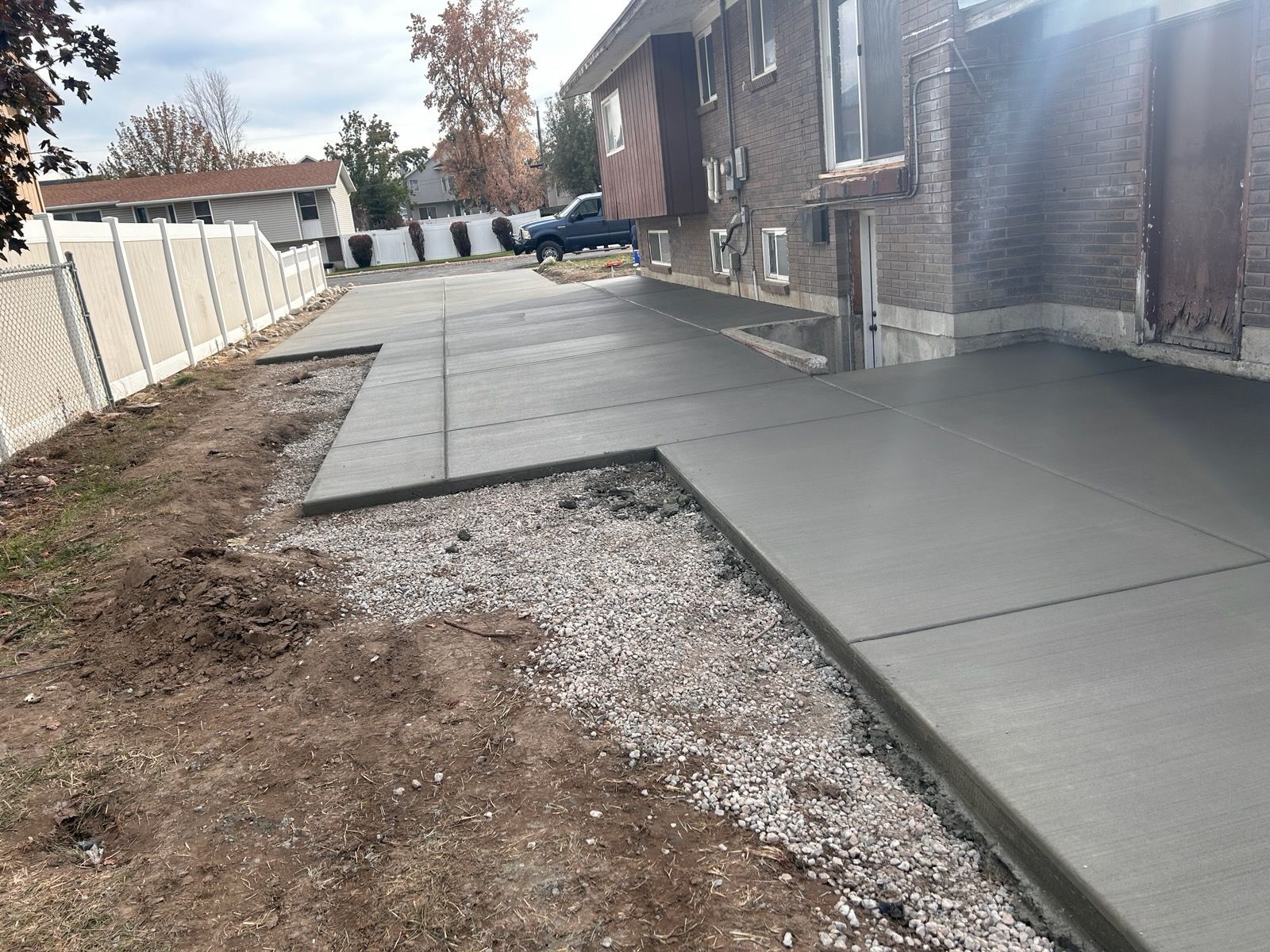 A newly poured concrete patio runs along the side of a brick house next to a gravel strip and a white fence.