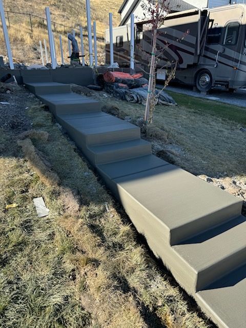 Concrete stairs recently poured on a grassy slope next to a construction site with an RV and fence posts in the background.