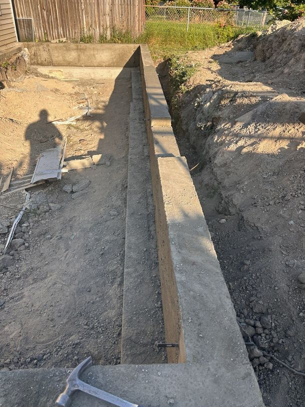 A concrete foundation wall under construction in a dirt excavation site, with a hammer resting on the foreground edge.