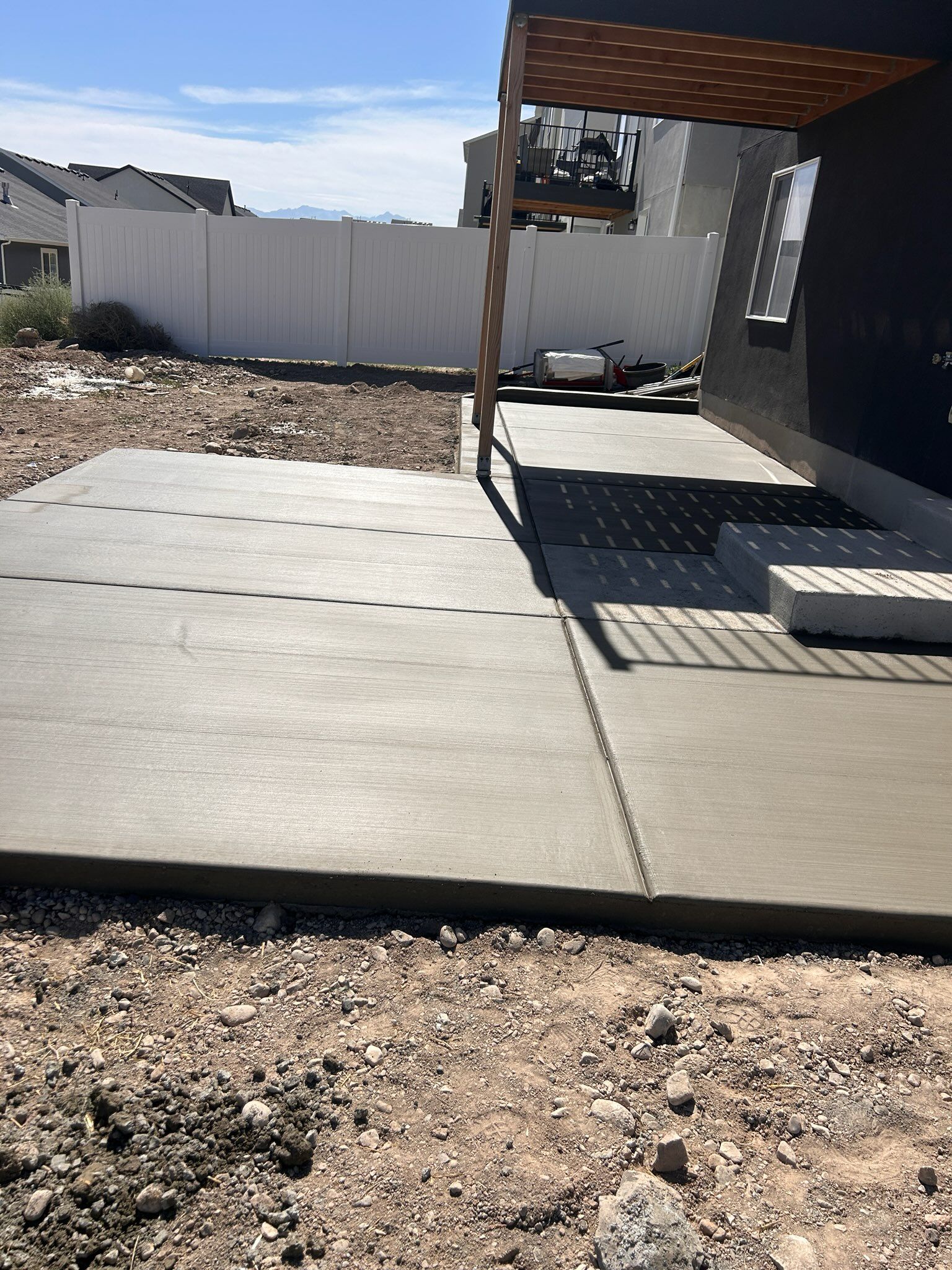A freshly poured concrete patio slab outside a modern home under a pergola, set in a yard with gravel and a white fence.