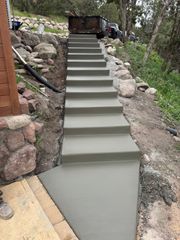 A set of newly poured, uniform concrete steps ascending a sloped, rocky hillside next to a wood-sided building.
