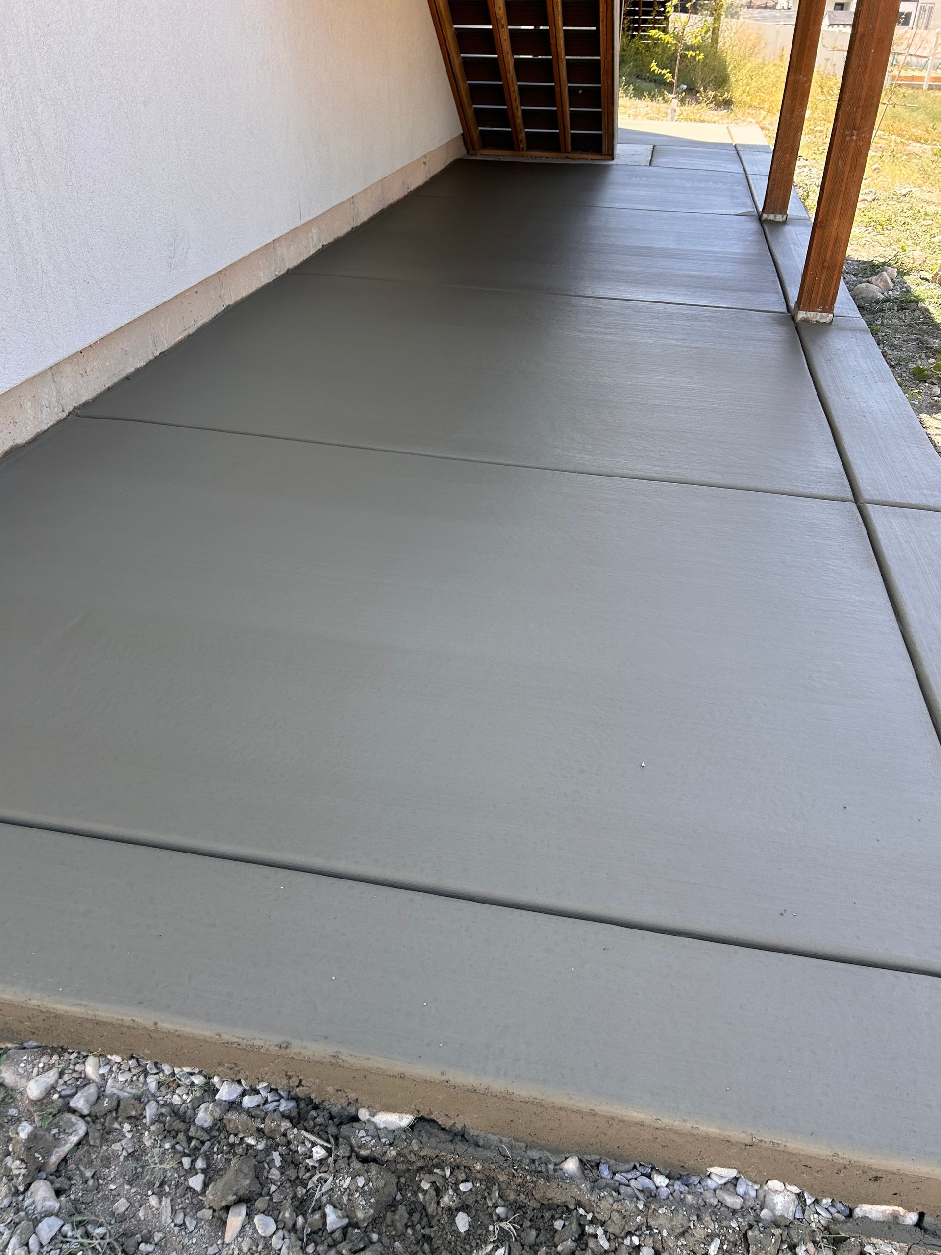 A freshly poured grey concrete walkway running alongside a building wall with wooden deck stairs above.
