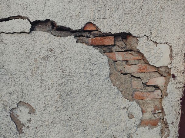 A section of an old, weathered wall with large patches of gray stucco crumbling to reveal red bricks beneath.