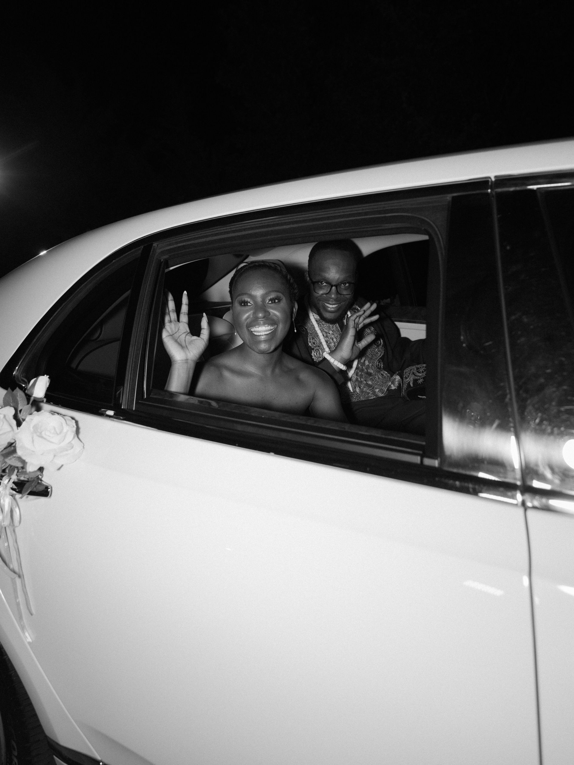 Two people smiling and waving from the back seat of a white car at night