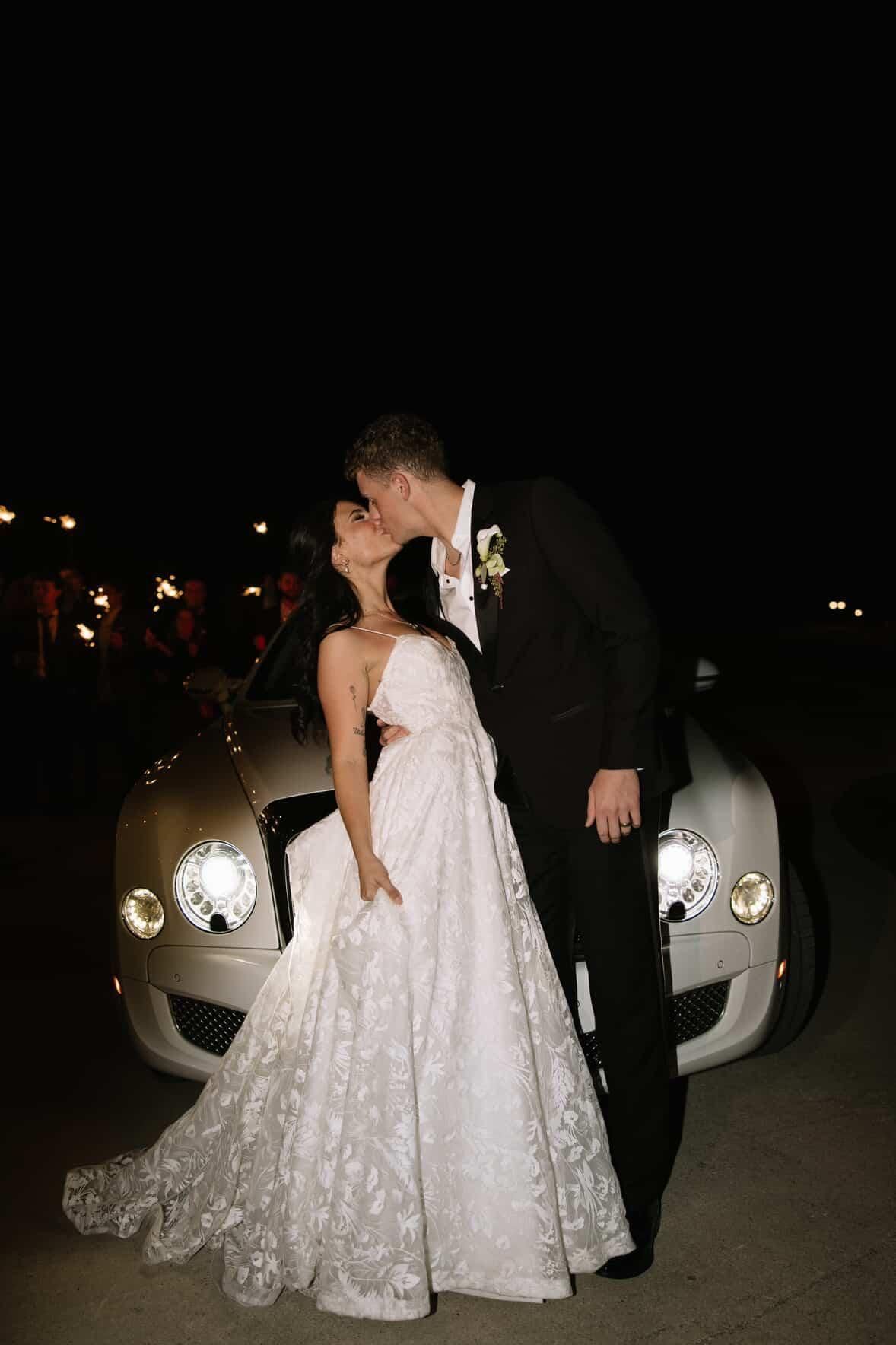 A bride and groom in formal wedding attire kiss in front of a white luxury car at night.