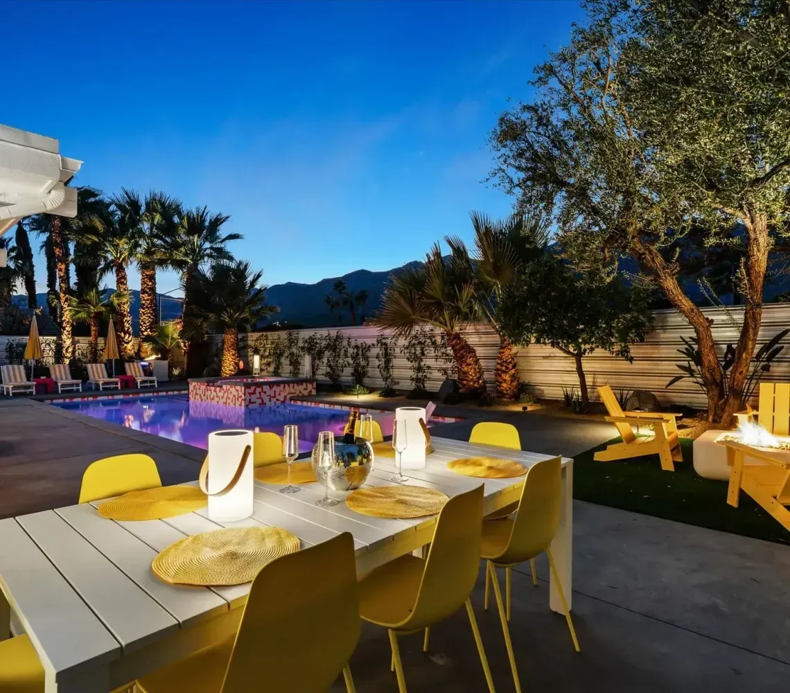 Outdoor patio with dining table, yellow chairs, and lounge seating at sunset under trees and blue sky