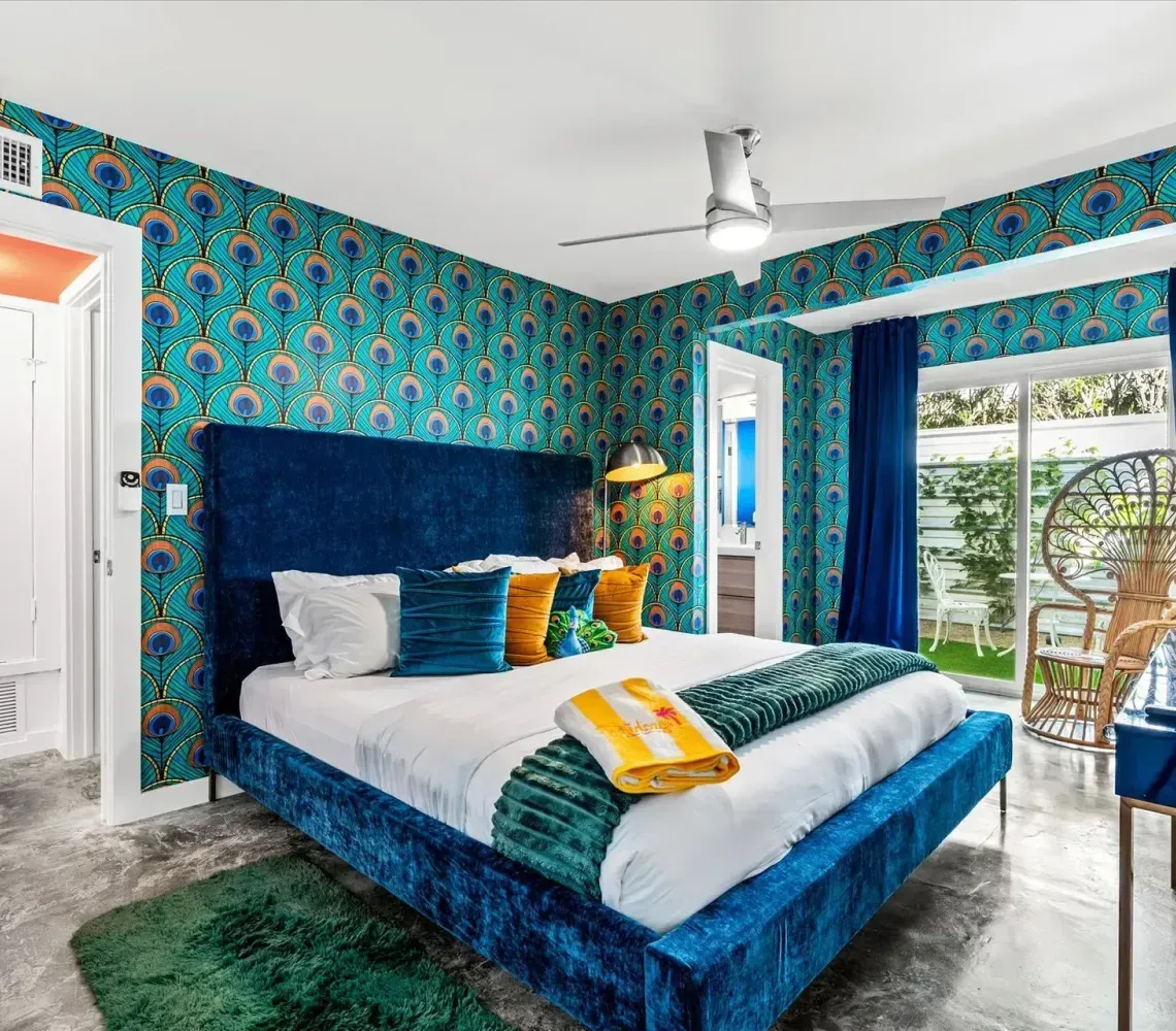 Colorful bedroom with a blue bed, patterned wallpaper, open balcony doors, and a ceiling fan