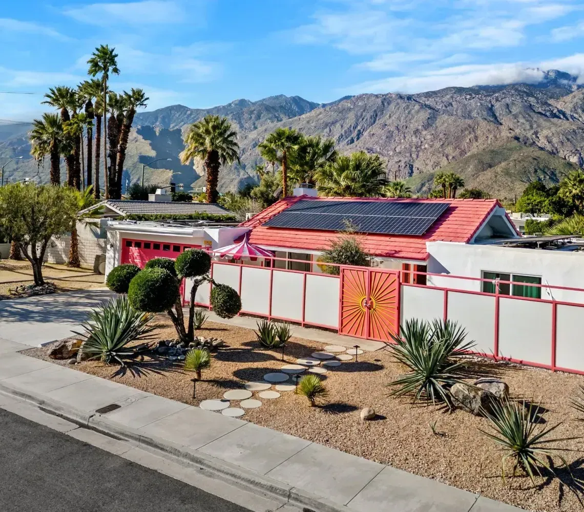 Desert home with pink roof and white walls, surrounded by palms and desert landscaping, with mountains in the background