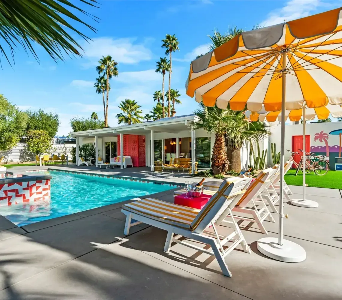 Poolside patio with orange-and-white umbrella, lounge chairs, and a turquoise swimming pool in a sunny resort setting
