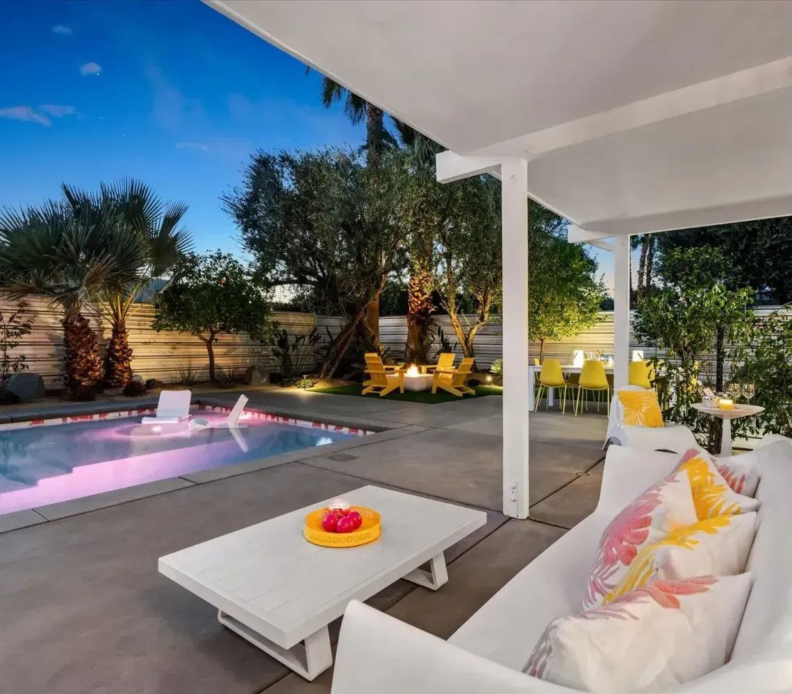 Covered patio with white sofa, coffee table, and lounge chairs beside a lit backyard pool at dusk