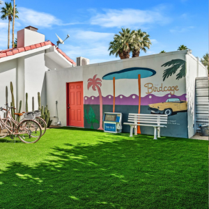 Colorful mural on a white building with a red door, bench, bicycle, palm trees, and bright green lawn.