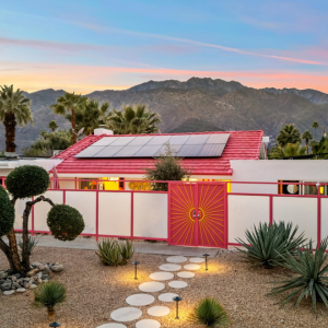 Desert home with red roof, solar panels, and a white wall, framed by mountains at sunset.