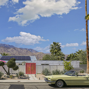 Palm-lined desert home with solar panels, mountains, and a green vintage convertible in front yard