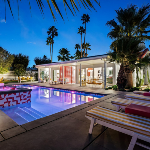 Modern luxury home with lit pool, patio lounge chairs, and palm trees at dusk