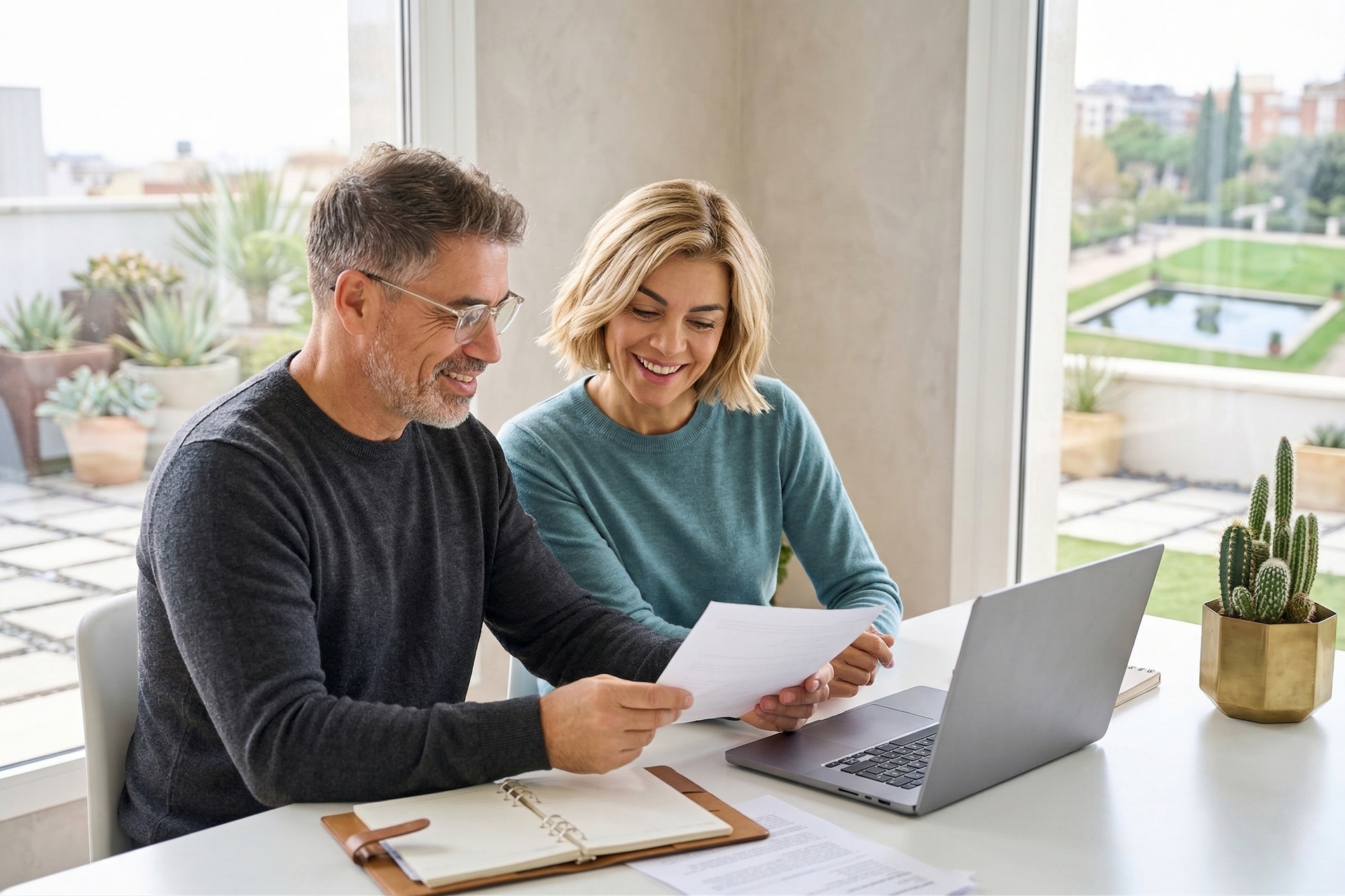 Two coworkers reviewing papers beside a laptop at a bright office desk, smiling