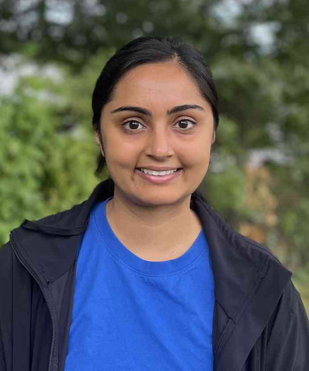 Person smiling outdoors in a blue shirt and dark jacket, with a blurred green background.
