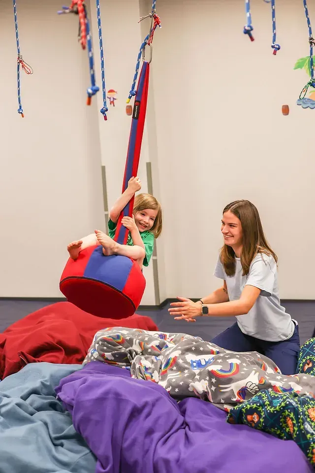 Child swinging on a red sensory swing while another child and an adult sit nearby in a playroom