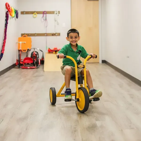 Child riding a yellow tricycle indoors, smiling in a bright hallway.