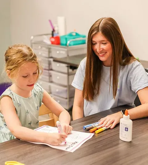 Child drawing at a table while an adult smiles beside her, with markers and craft supplies nearby.