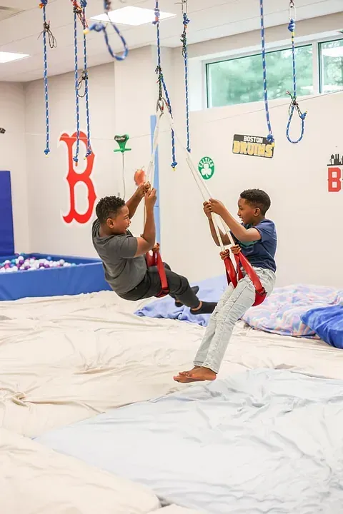 Two children swinging on ropes in an indoor play area with white mats and hanging toys.
