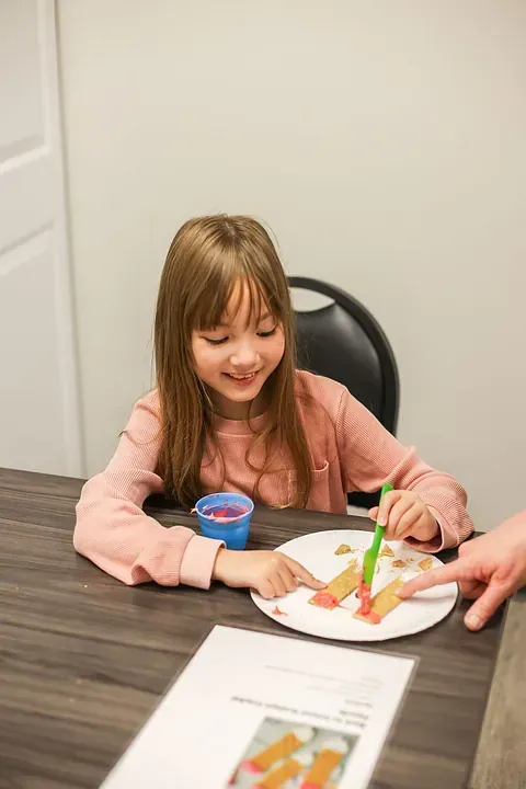 Child smiling at a table, eating cake with a fork while an adult steadies the plate.