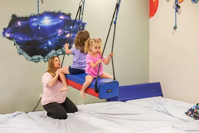 Two children on swings in a white-walled sensory room, with a caregiver kneeling beside them.