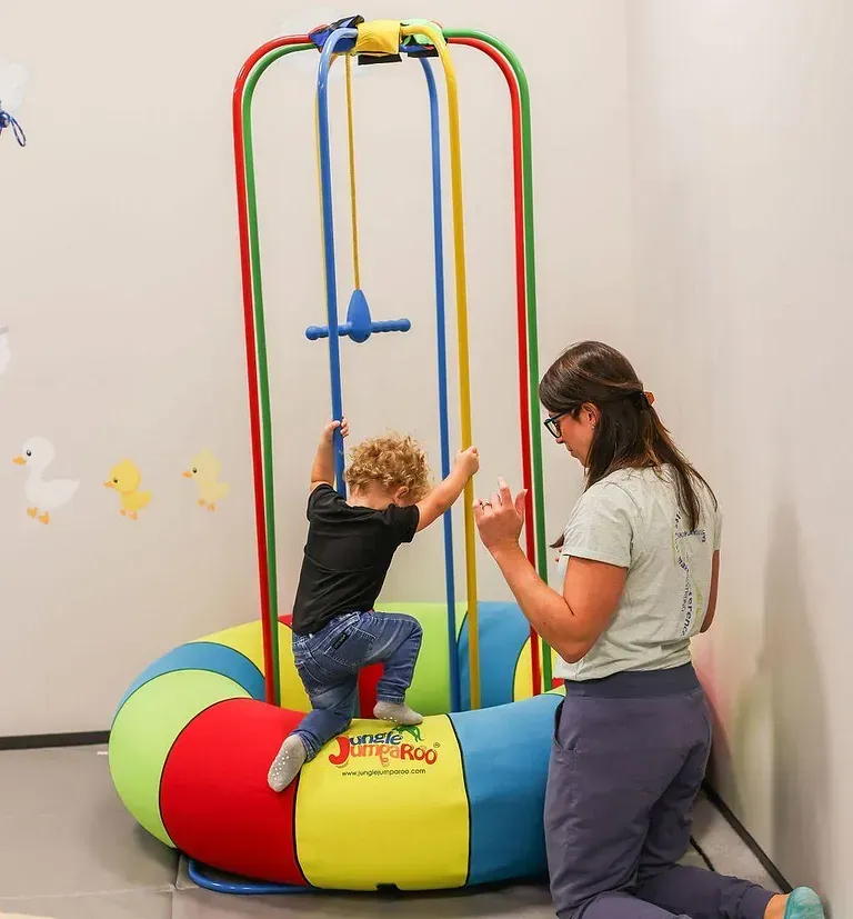 Child on colorful indoor swing with an adult nearby in a playroom