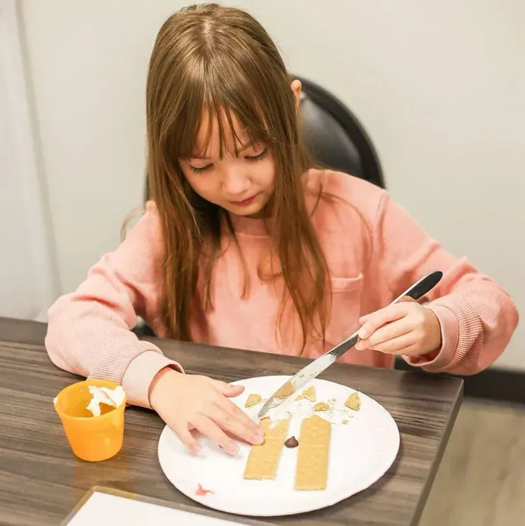 Child using a fork to arrange food on a white plate at a table, with an orange cup nearby