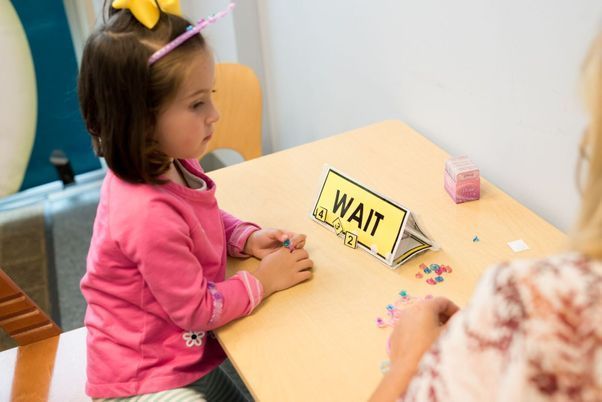 Child in a pink shirt sits at a table with a “WAIT” sign and small colorful pieces.