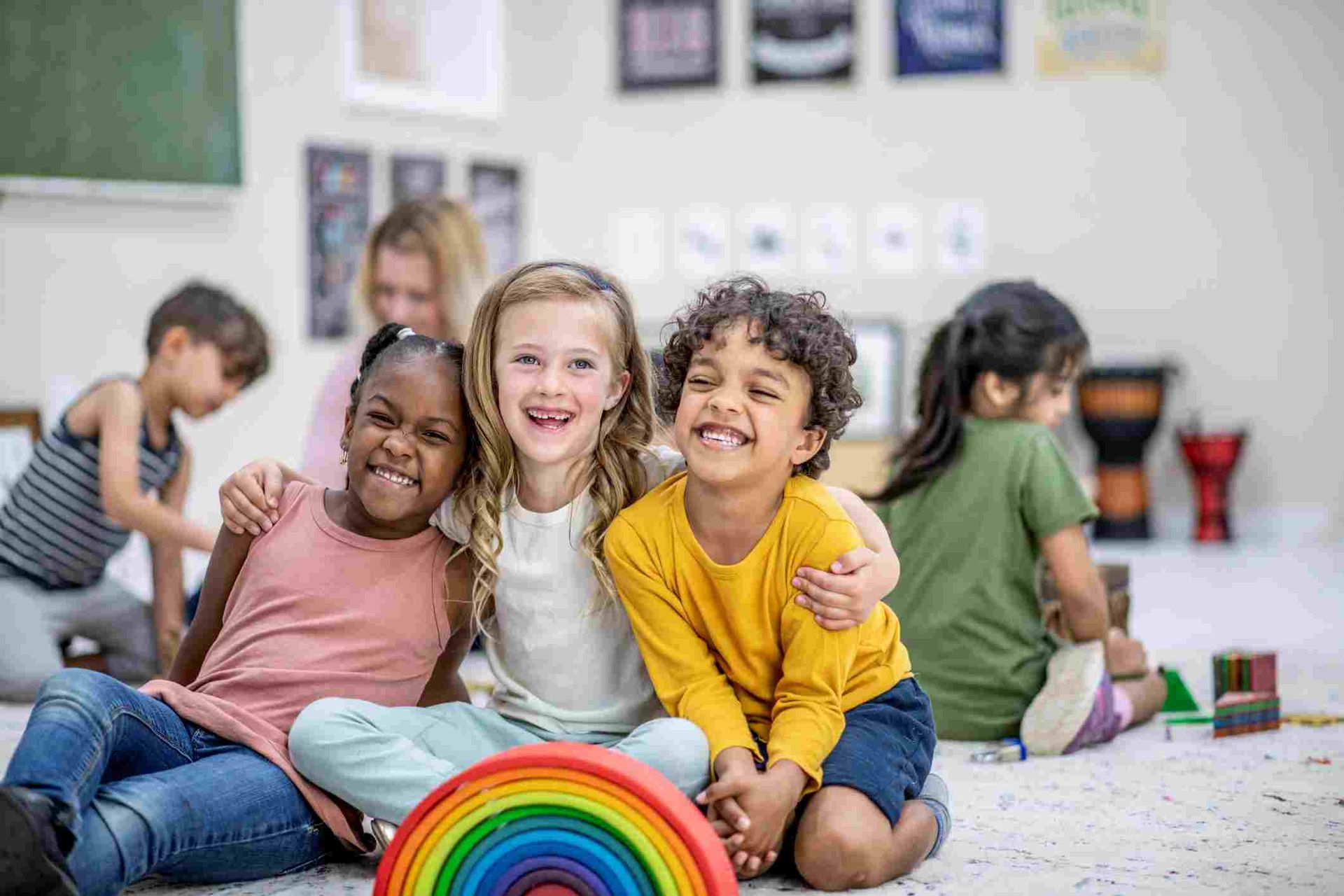 Children playing with toys on a classroom floor, smiling and sitting together in a bright play area.