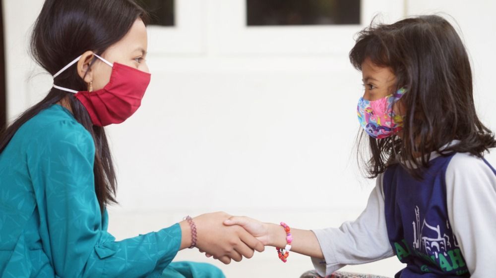 Two children wearing face masks hold hands while seated across from each other indoors