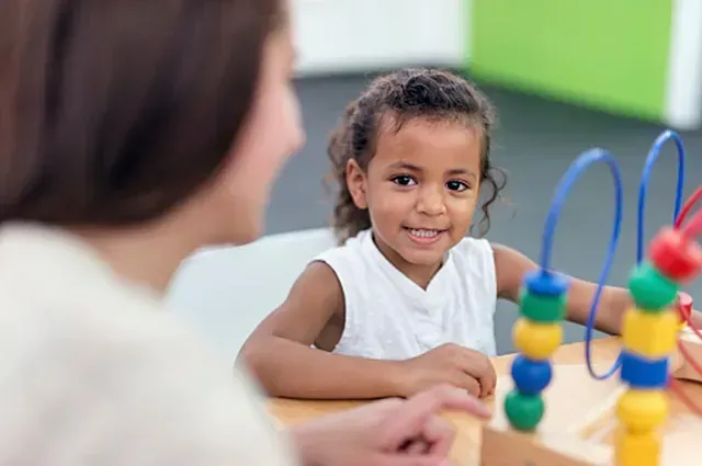 Child smiling at a classroom table with colorful bead loops, facing an adult nearby