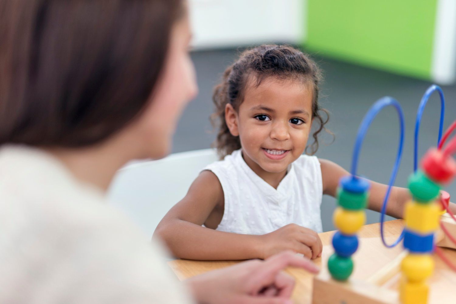 Child smiling at a bead maze table in a classroom, with an adult nearby
