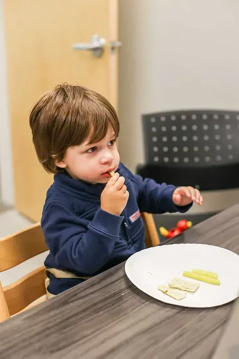Toddler eating a snack at a table in a classroom, with a plate of fruit in front of them