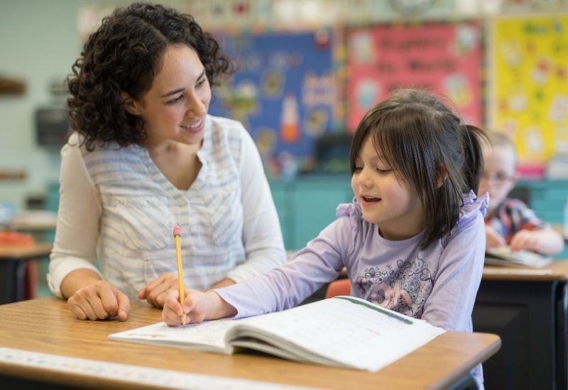 Teacher helping a child write at a classroom desk