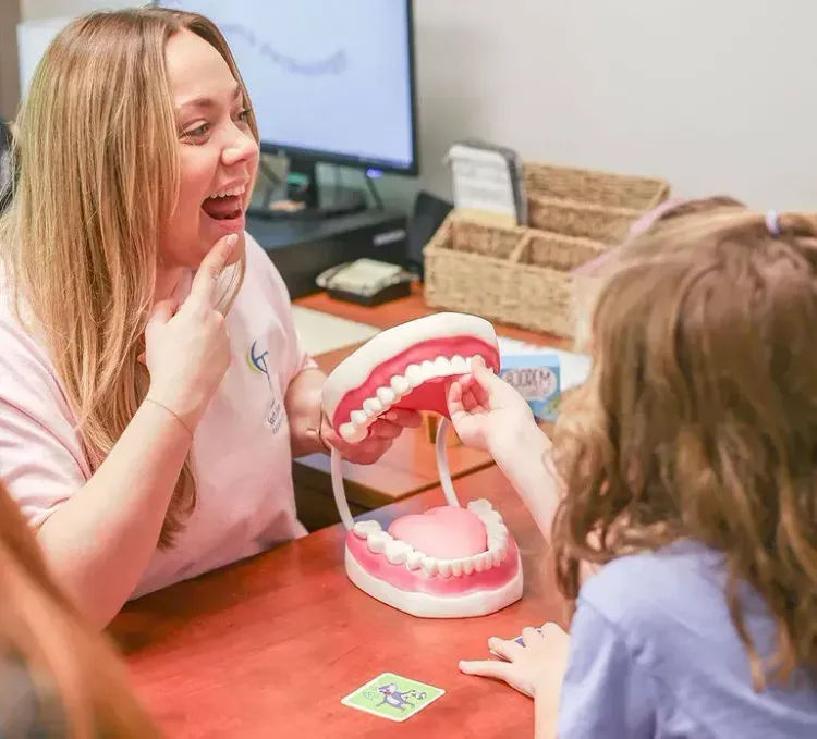 Child playing dentist with a model teeth set at a table, while another smiles nearby.