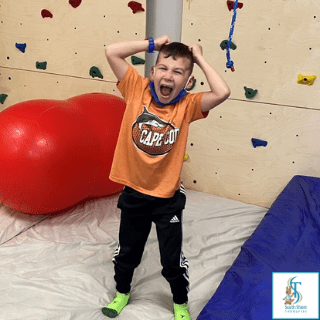 Child cheering on an indoor climbing wall, wearing an orange shirt, with a large red ball nearby.