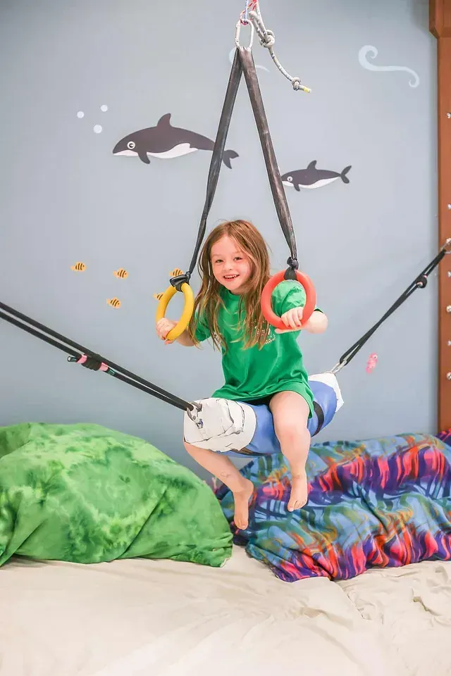 Child on hanging gym rings in a playful indoor ocean-themed room, smiling on colorful cushions.
