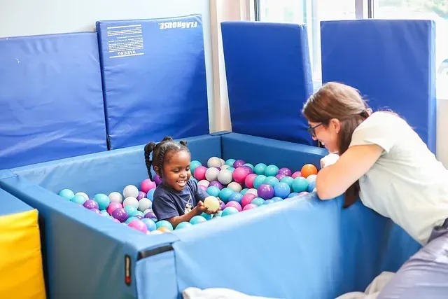 Child in a blue ball pit smiling at a caregiver leaning over the edge