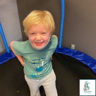 Smiling child standing on a trampoline indoors, wearing a green shirt and light pants.
