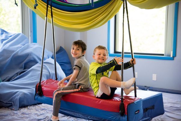 Two children sit on a blue indoor swing with colorful draped fabric in a playroom.