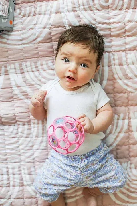Baby lying on a quilt, holding a pink toy ball and looking up with a curious expression