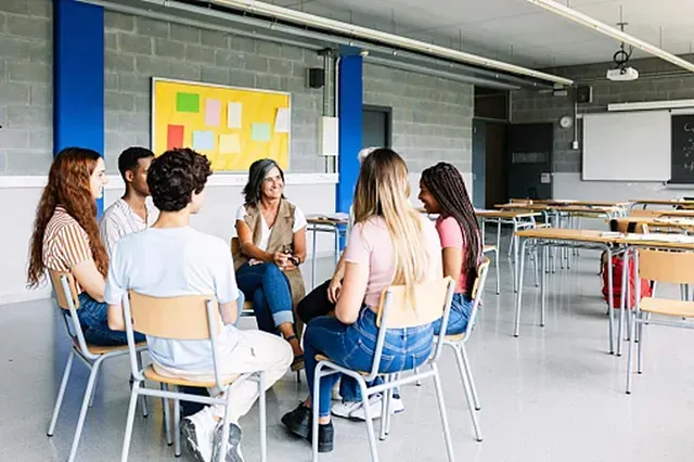 People seated in a circle, listening to a speaker in a bright classroom or meeting room