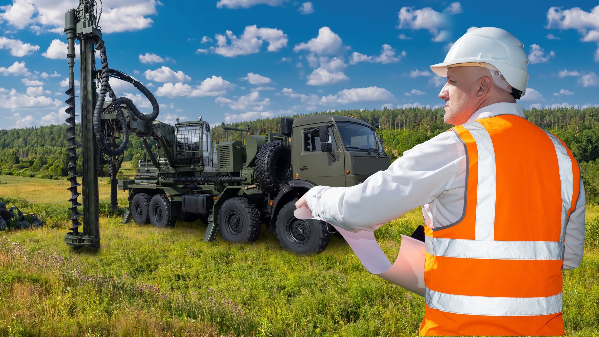 Worker in orange vest and hard hat observing drilling rig and truck in a grassy field under blue sky