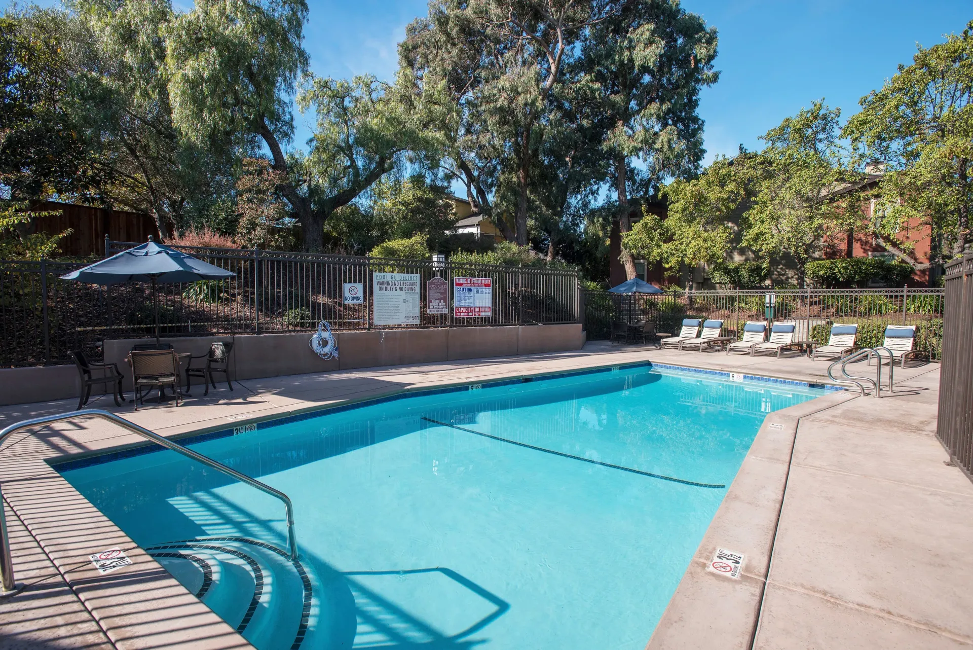 Outdoor apartment community pool with lounge chairs, tables, umbrellas, and surrounding trees at Sterling Heights apartments in Benicia, CA.